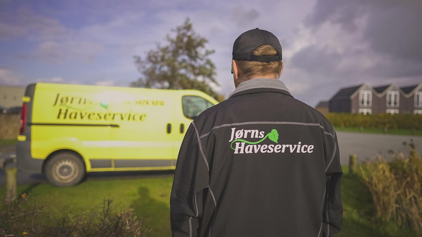 En mand i en sort jakke står med ryggen til en gul varevogn med teksten "Jarn's Haveservice" på.