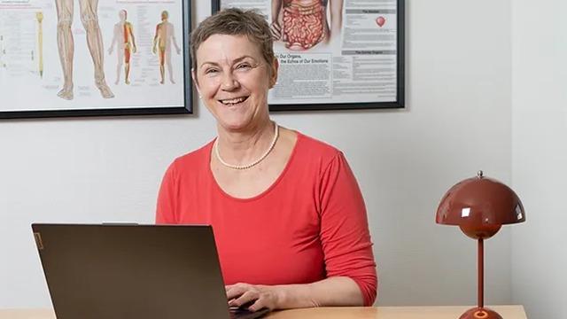 En smilende kvinde i rød bluse sidder ved et skrivebord med en bærbar computer og en lampe i et kontormiljø.
