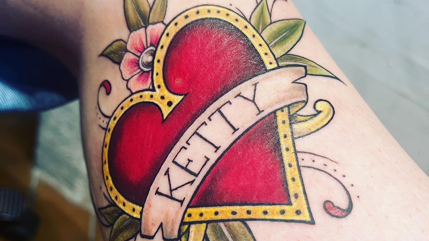 Farverig tatovering af et hjerte med navnet "KETTY" og blomsterdekorationer omkring.