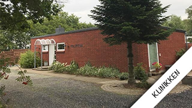 Rød murstensbygning med indgang og grønne planter foran, beliggende i et roligt område.