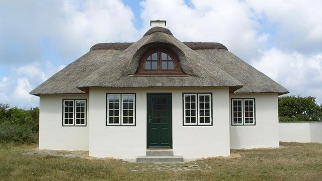Et charmerende hus med stråtag og grøn dør, omgivet af græs og en blå himmel med skyer.