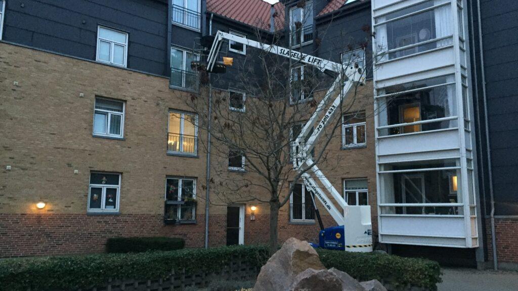 En lift står ved en bygning, mens en arbejder reparerer vinduer på første sal.