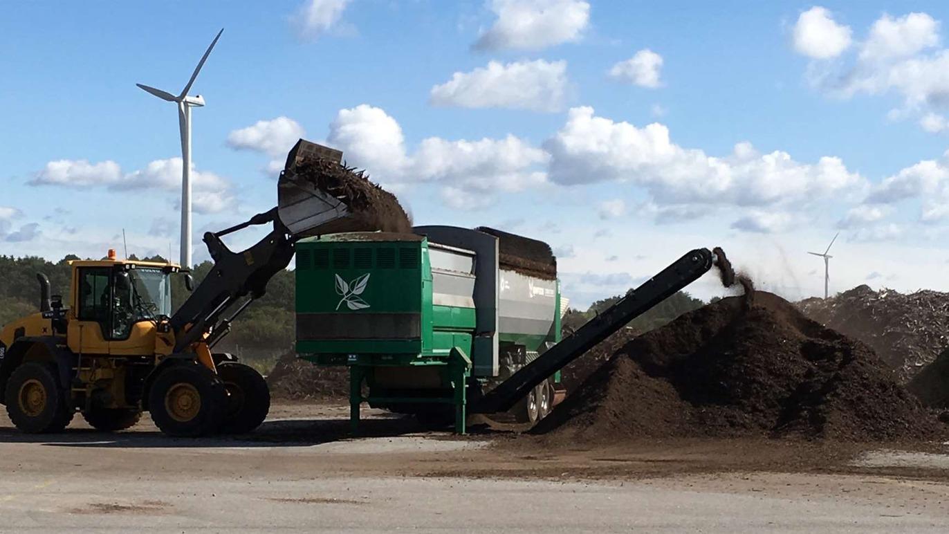 En gule traktor læsser kompost i en grøn maskine, mens der er vindmøller i baggrunden.
