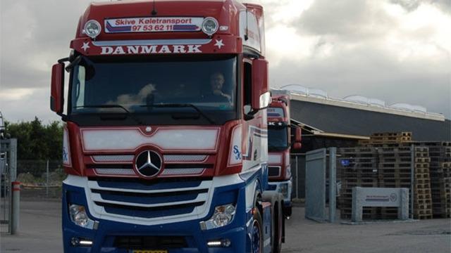 En stor lastbil med dansk flag og teksten "DANMARK" kører på en vej ved en lagerbygning.