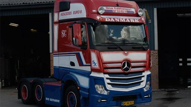 En farverig lastbil med dansk flag og logo, parkeret foran en garage.