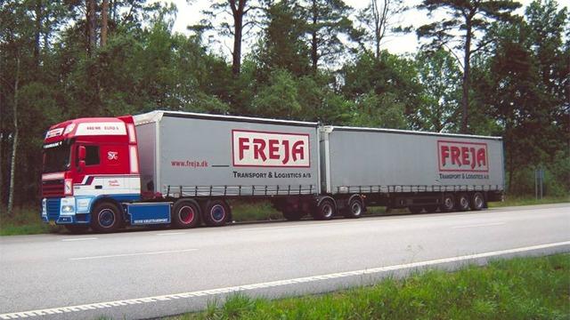 En stor lastbil med to trailere, der har FREJA-logoet, parkeret ved en vej omgivet af træer.
