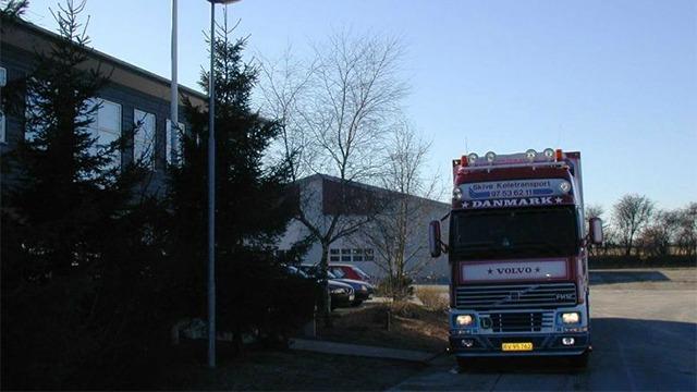En rød lastbil parkeret foran en bygning med træer i baggrunden.