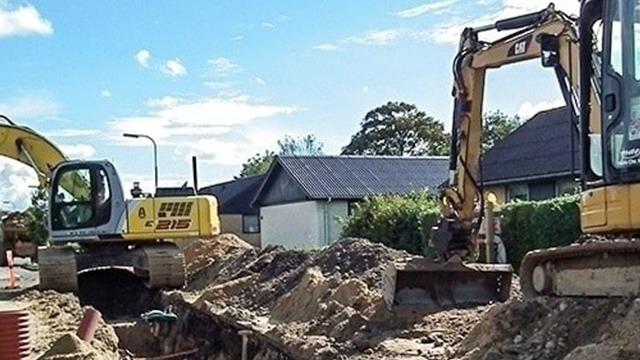 To store med gravemaskiner arbejder på en byggeplads med sand og grus under en blå himmel.