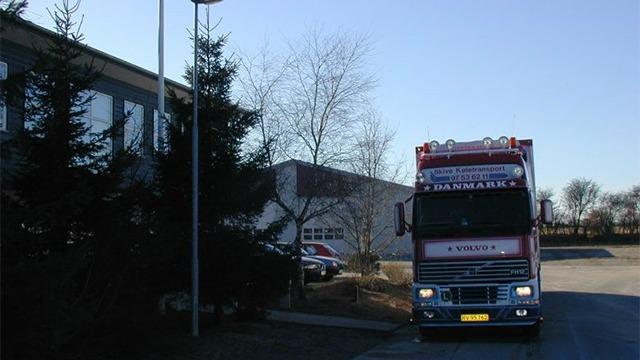 En lastbil parkeret foran en bygning med træer og gadebelysning i dagslys.