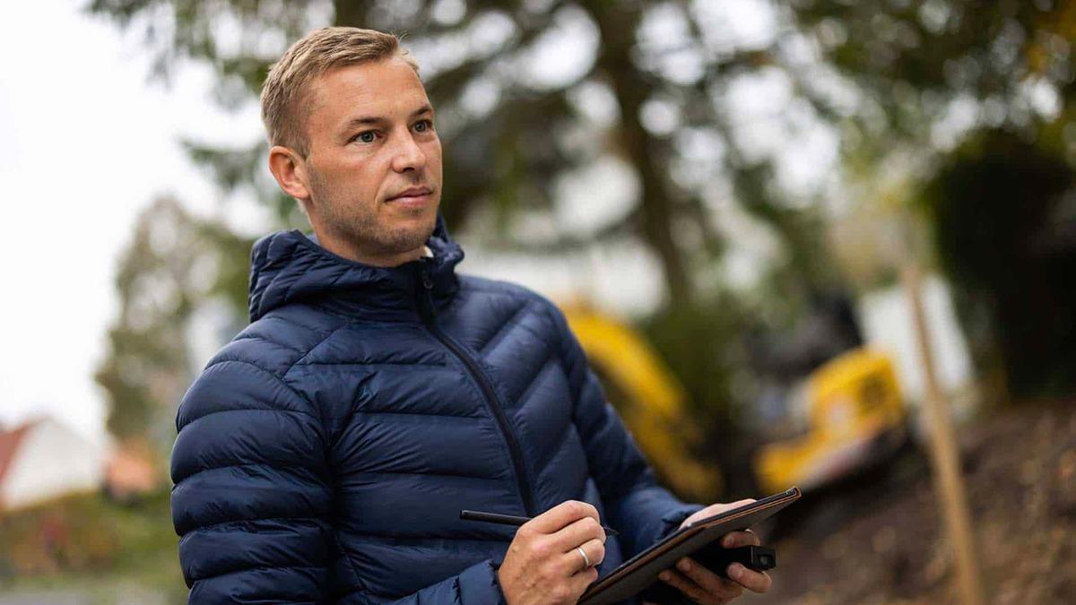 En mand i en blå jakke står med en tablet og noterer, mens han ser på en byggeplads i baggrunden.