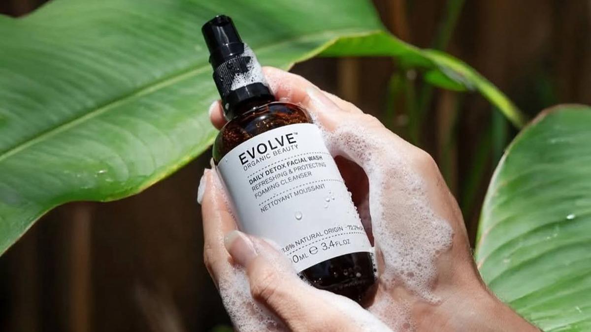 Hånd holder en flaske Evolve ansigtsspray med skum, omgivet af grønne planter.