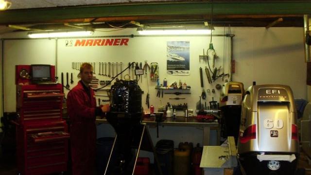 En mekaniker arbejder i et bådværksted med værktøj og motorer i baggrunden.