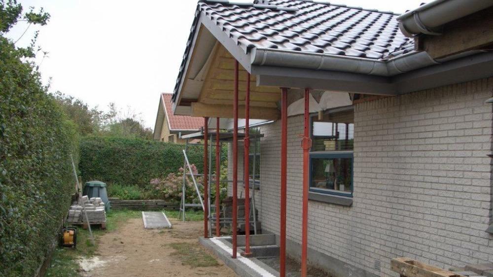 Hus med tagudhæng og støttepiller, omgivet af have og byggeplads.