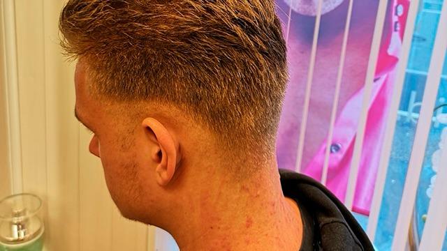 En ung mand med kort hår og en fade-frisure, der står ved et vindue med persienner.