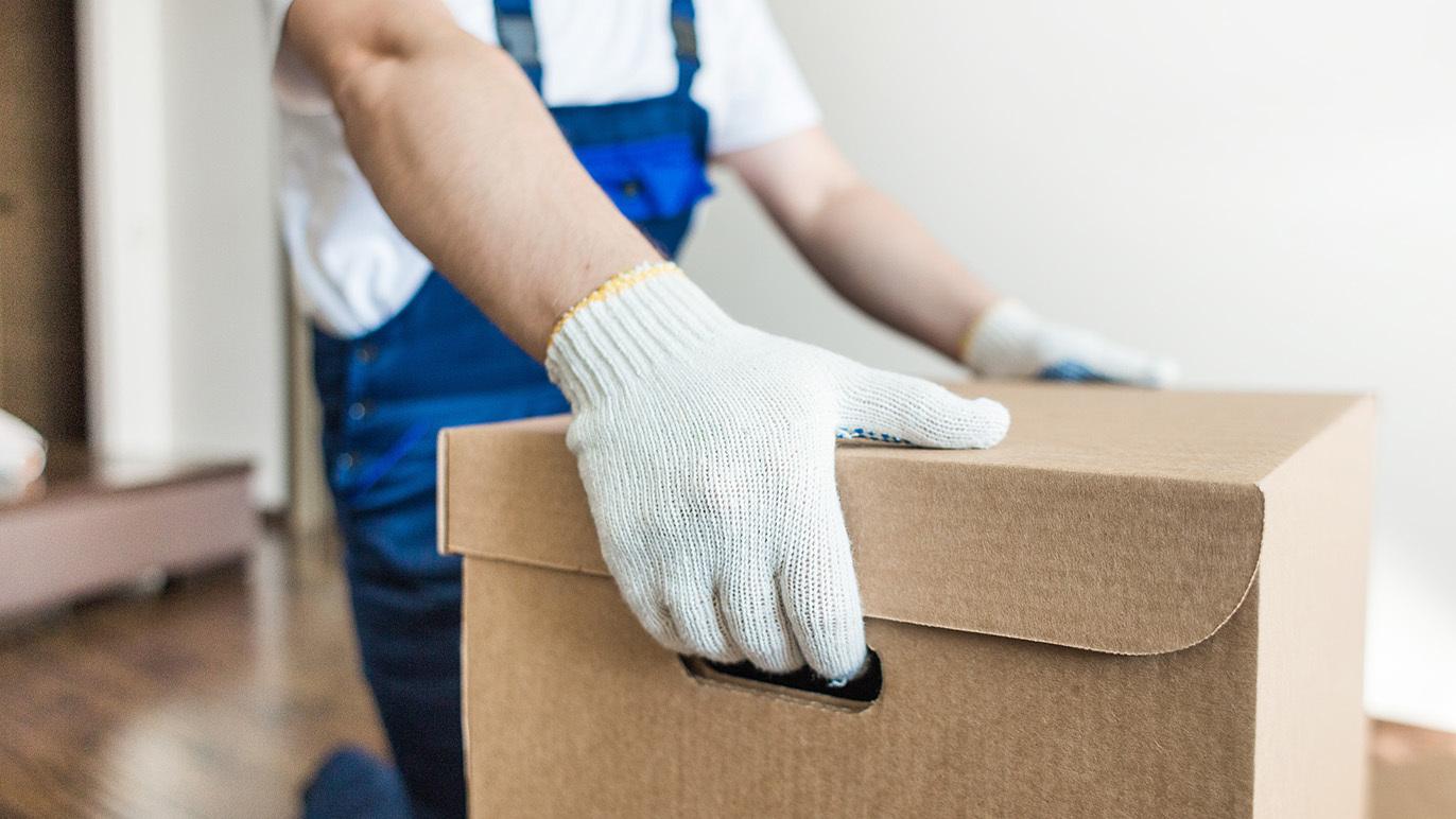 En person i arbejdstøj bærer en papkasse med handsker i et lyst rum.