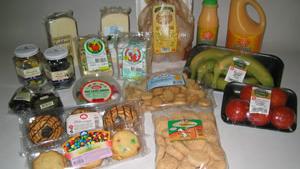 Billede af en indkøbskurv med forskellige fødevarer, herunder frugt, kager, ost, oliven og juice.