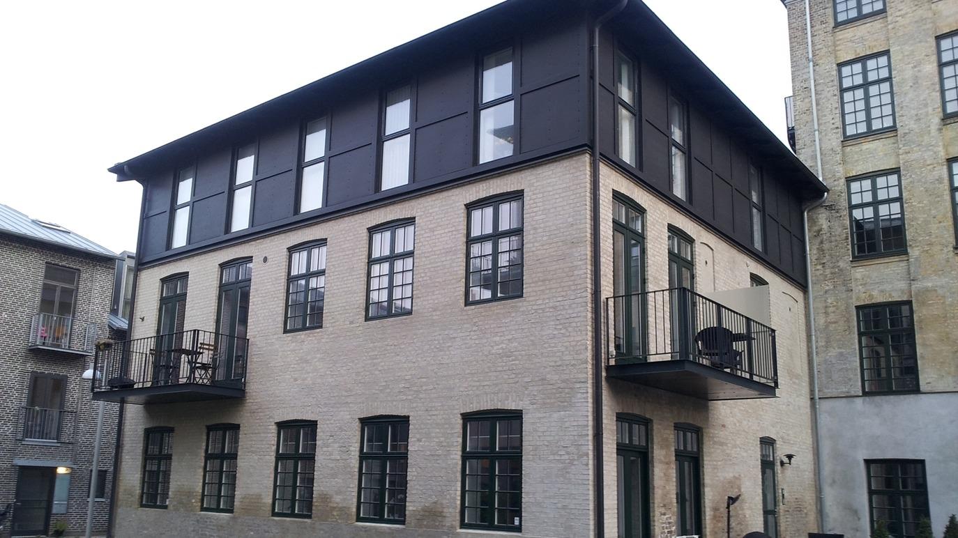 Moderne bygning med sorte vinduer og altaner, beliggende i en bymiljø med murstensbygninger.