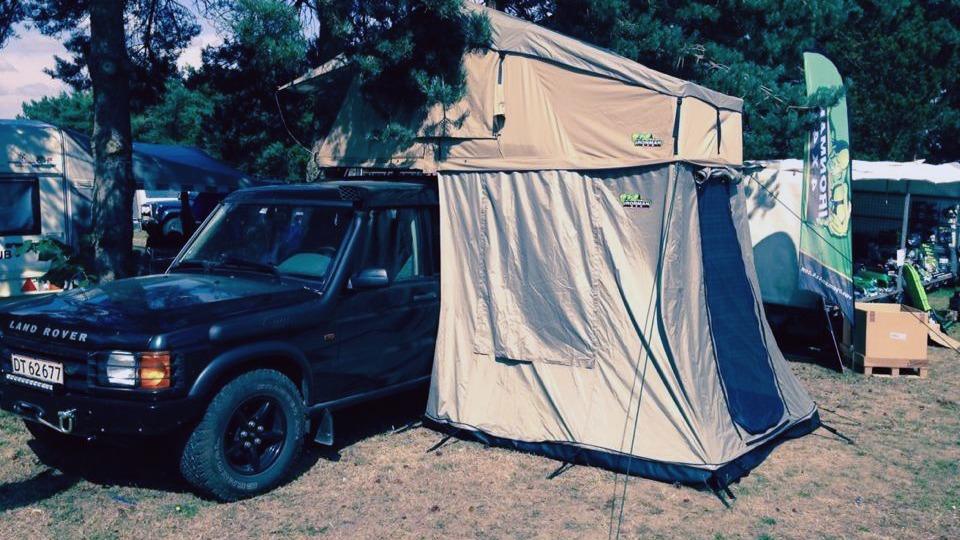 En sort bil med tagtelt og fortelt, parkeret på en campingplads omgivet af træer og telte.