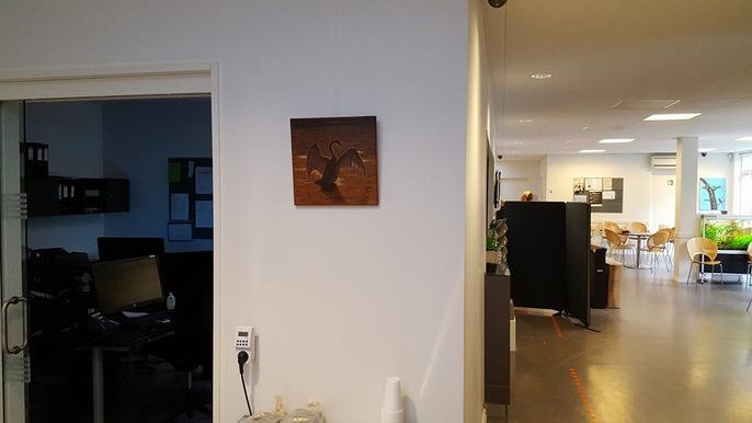 Billede af en væg med et maleri og et kontormiljø i baggrunden, der viser skriveborde og planter.