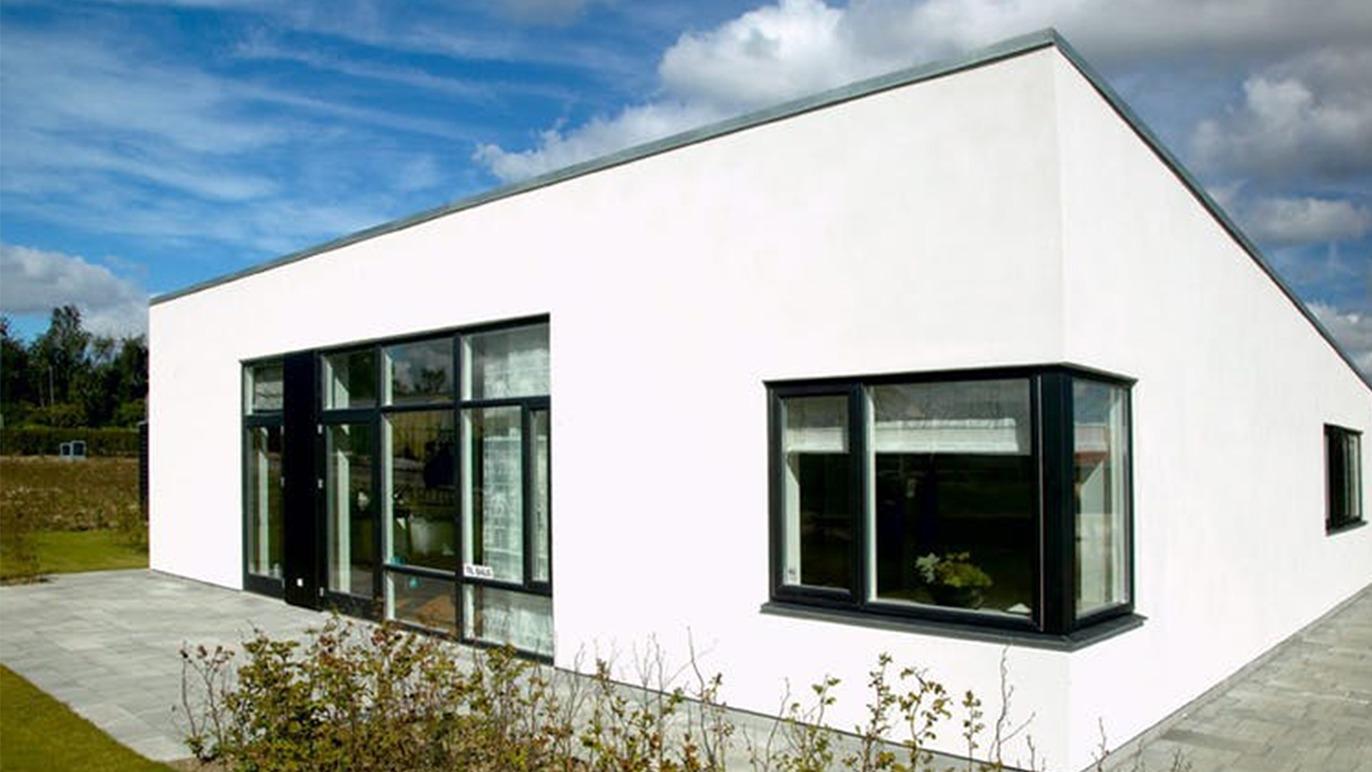 Moderne hvidt hus med store vinduer og fladt tag, omgivet af grønne områder og blå himmel.