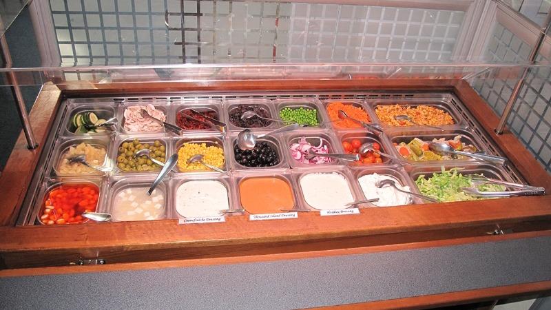 Et salatbar med forskellige toppings som grøntsager, saucer og nødder i metalbeholdere.