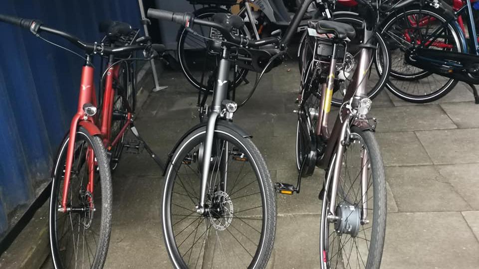 Tre cykler parkeret ved en væg, herunder en rød, en sort og en brun cykel.