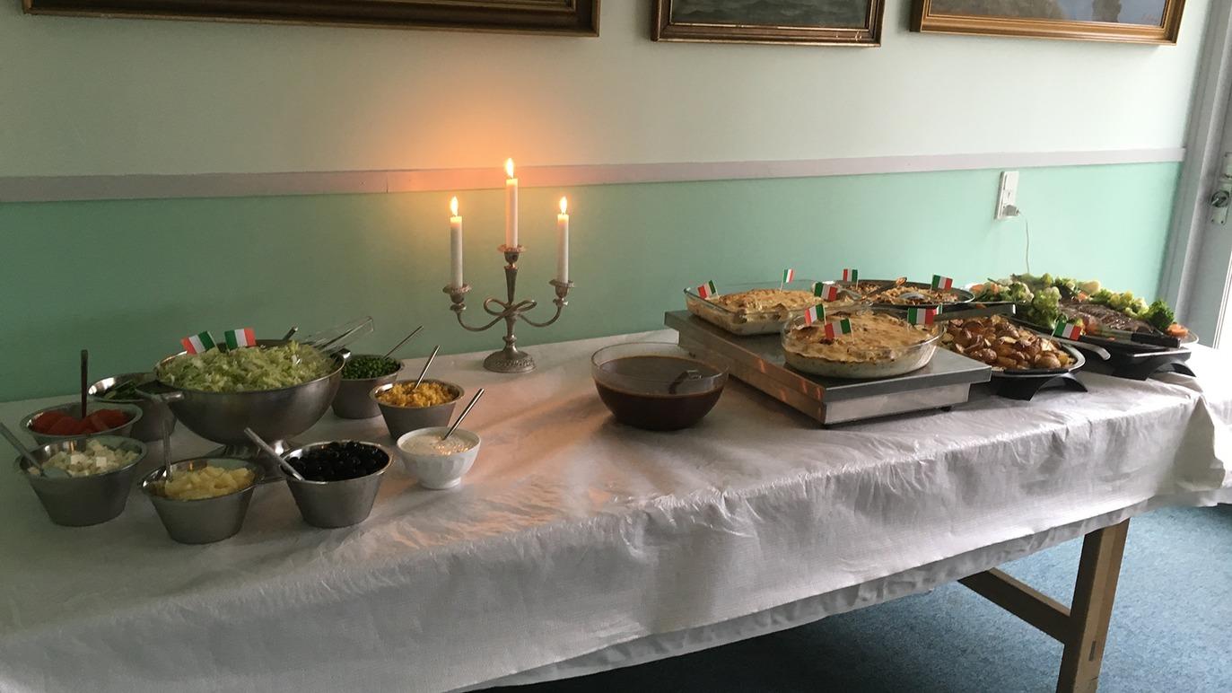 Et festligt buffetbord med salater, kager og snacks, oplyst af stearinlys i en hyggelig atmosfære.
