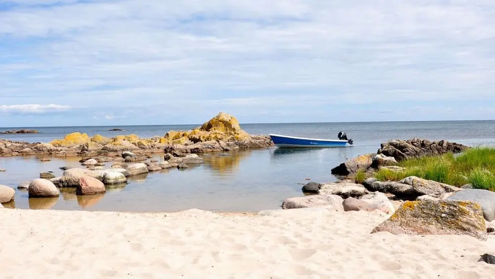 En båd ligger ved kysten nær en stenstrand med klipper og græs under en blå himmel.