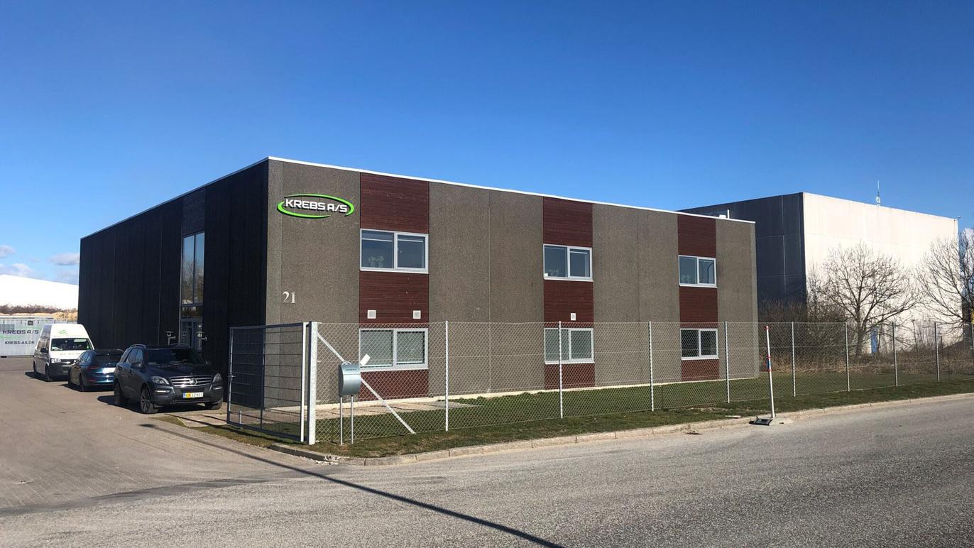 Moderne erhvervsbygning med grå facade og vinduer, beliggende ved en vej med parkerede biler.