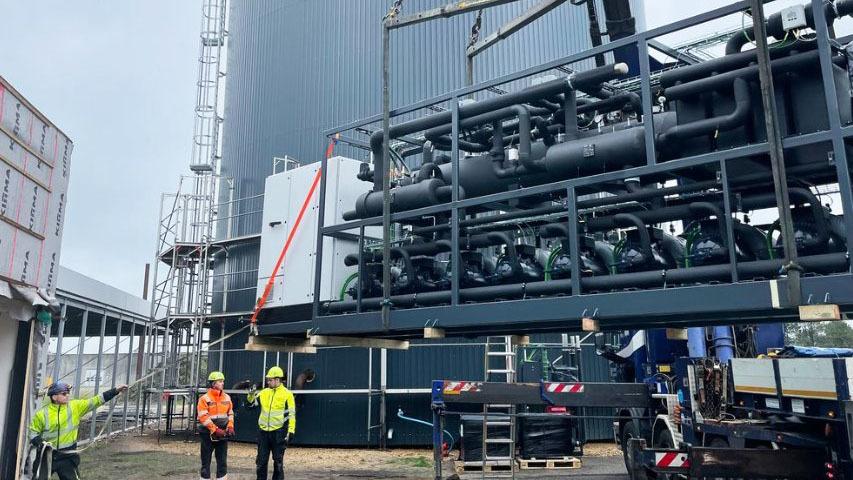 Arbejdsplads med to personer i sikkerhedstøj, der overvåger en kran, der løfter rørinstallationer ved en bygning.