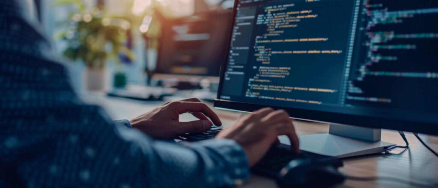 En person skriver kode på en computer med programmeringsskærm i et moderne kontormiljø.