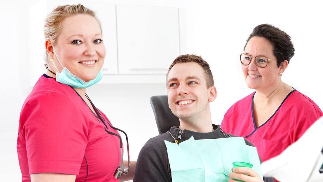 To tandlæger i pink uniformer smiler til en glad patient i tandlægestolen med en tandlægeassistent.