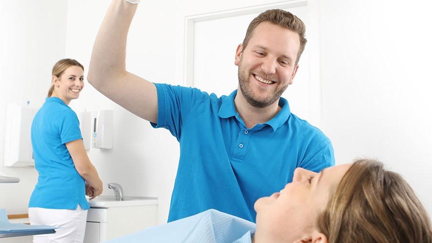 En tandlæge smiler, mens han holder en tandbørste over en patient, der ligger i tandlægestolen. En sygeplejerske står i baggrunden.