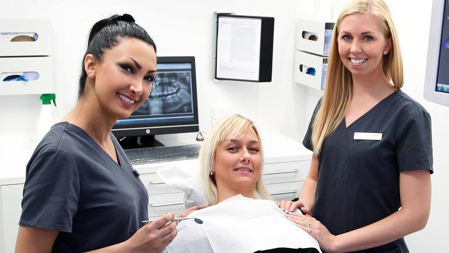 To kvinder i tandlægeklinikken smiler, mens de står ved en patient, der sidder i tandlægestolen.
