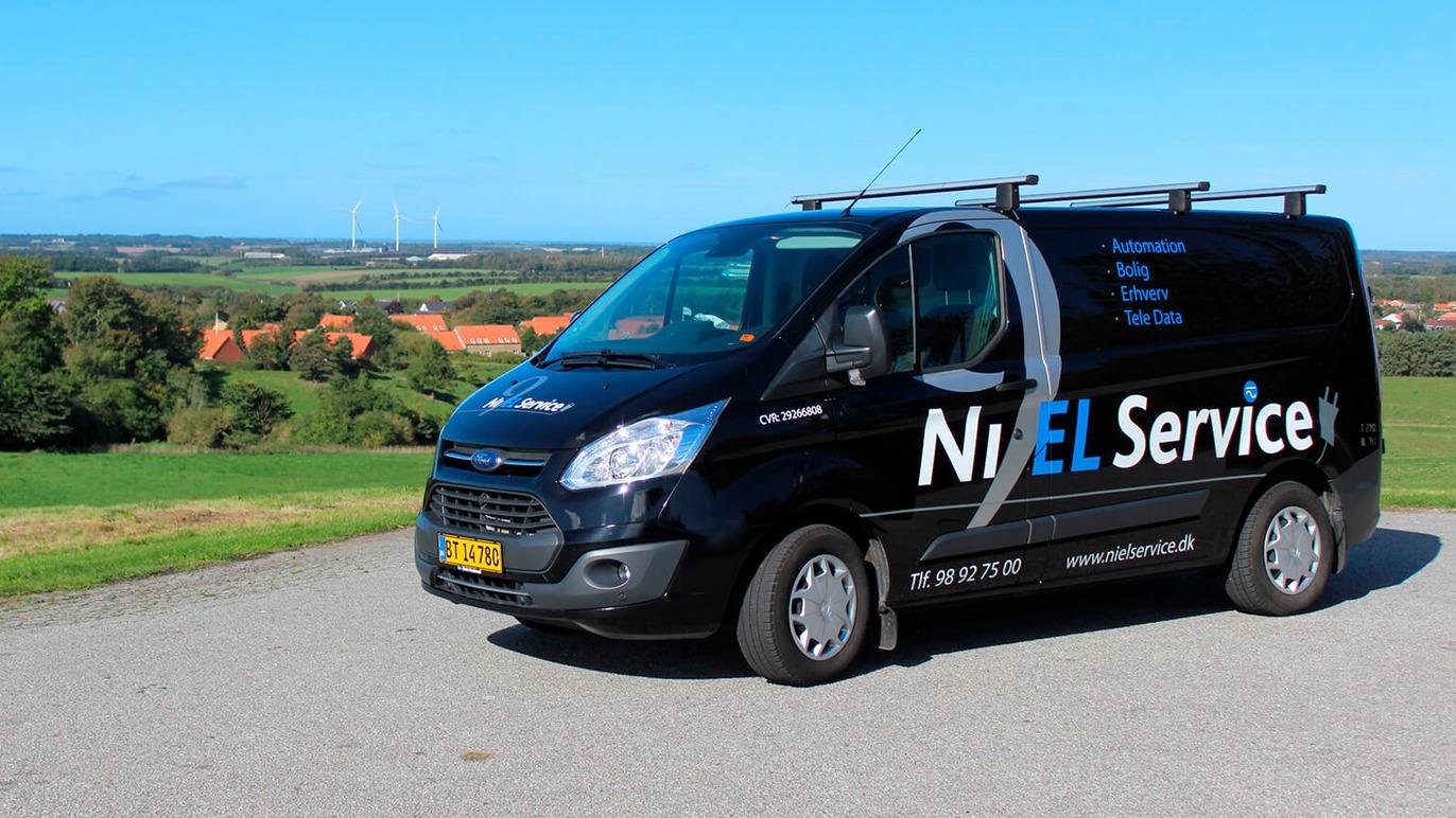 En sort varevogn med teksten "NixEL Service" parkeret på en vej med grønne marker og landsby i baggrunden.