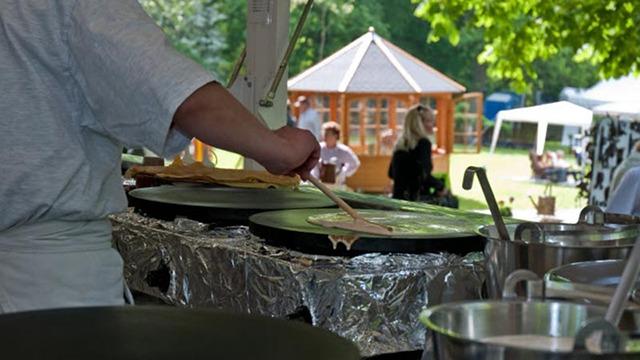 En person laver pandekager på en stor pande ved en udendørs festival med telte i baggrunden.