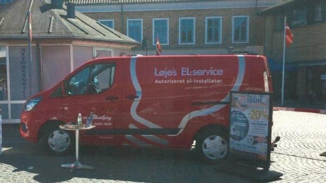 Rød varevogn med teksten "Løjes EL-service" parkeret ved en bygning med flag og et informationsskilt.