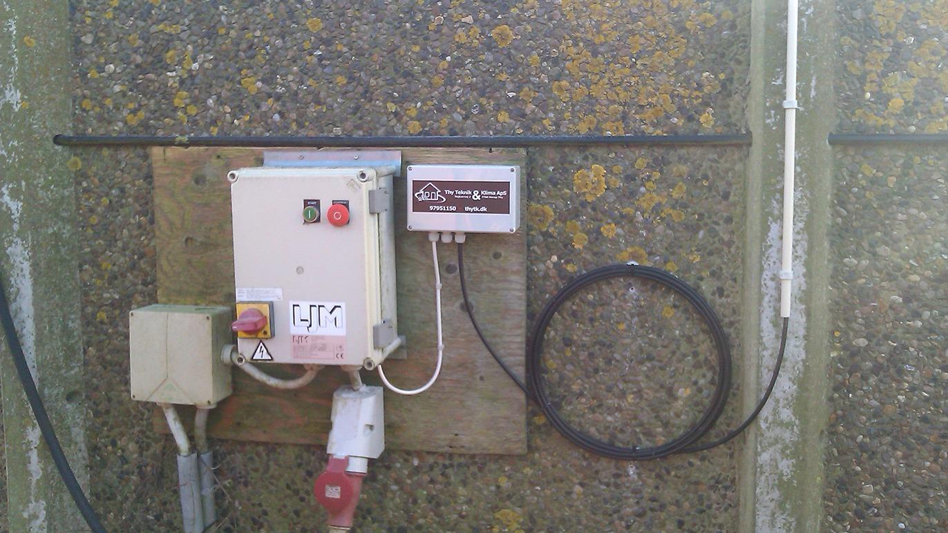 Boks med elektrisk kontrolpanel og tilslutninger på en væg, omgivet af mos og alger.