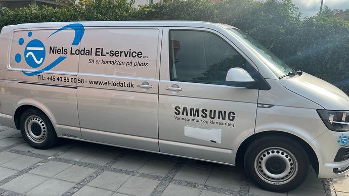 En sølvfarvet varevogn med teksten "Niels Lodal EL-service" og Samsung-logoet på siden, parkeret på en flisebelagt overflade.
