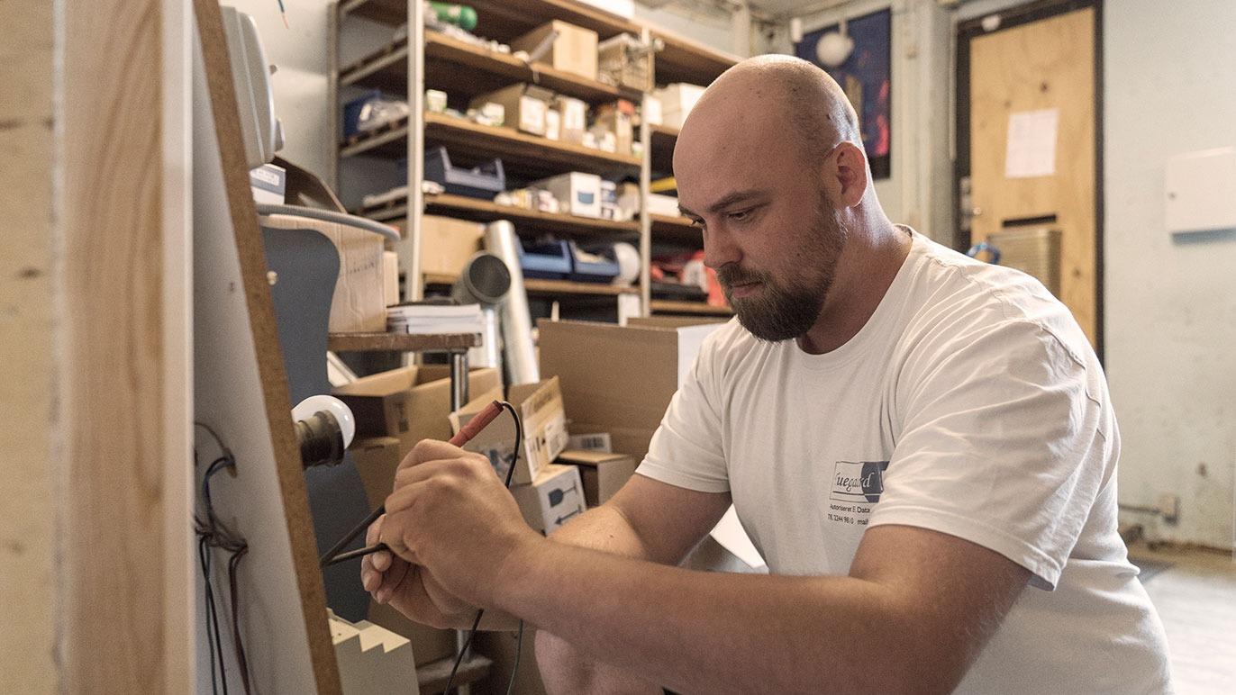 En mand sidder på gulvet og arbejder koncentreret med værktøj i et værksted fyldt med kasser og materialer.