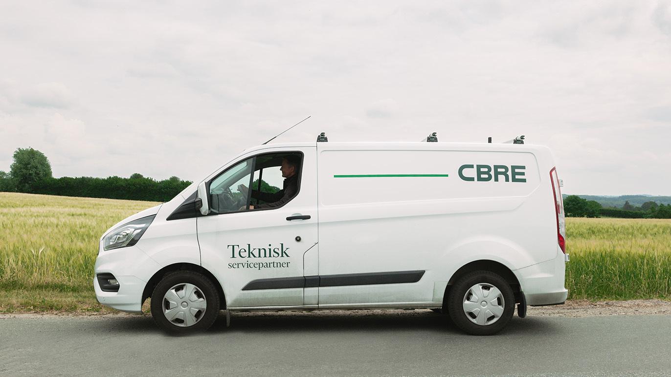 En hvid varevogn med teksten "CBRE" og "Teknisk service" parkeret ved en mark.