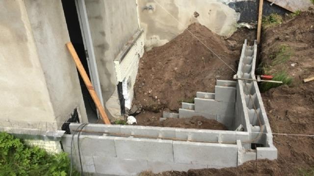 Byggeri af en kælder med betonblokke og jordhøje ved en husvæg.