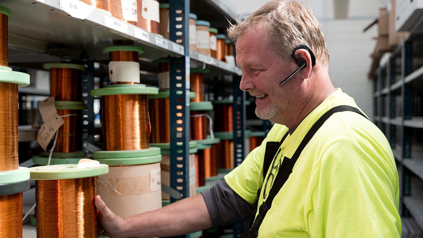 En mand i gul sikkerhedstrøje står ved en hylde med spoler af kobbertråd og smiler, mens han bruger et headset.