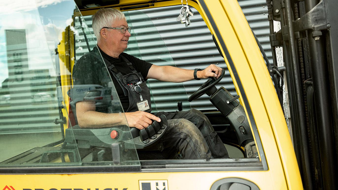 En mand sidder i en gaffeltruck og styrer den, iført arbejdstøj og sikkerhedsudstyr.