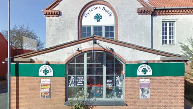 Butikslokale med skiltet "Dyrenes Butik" og vinduer, der viser dyreforsyninger og plakater.