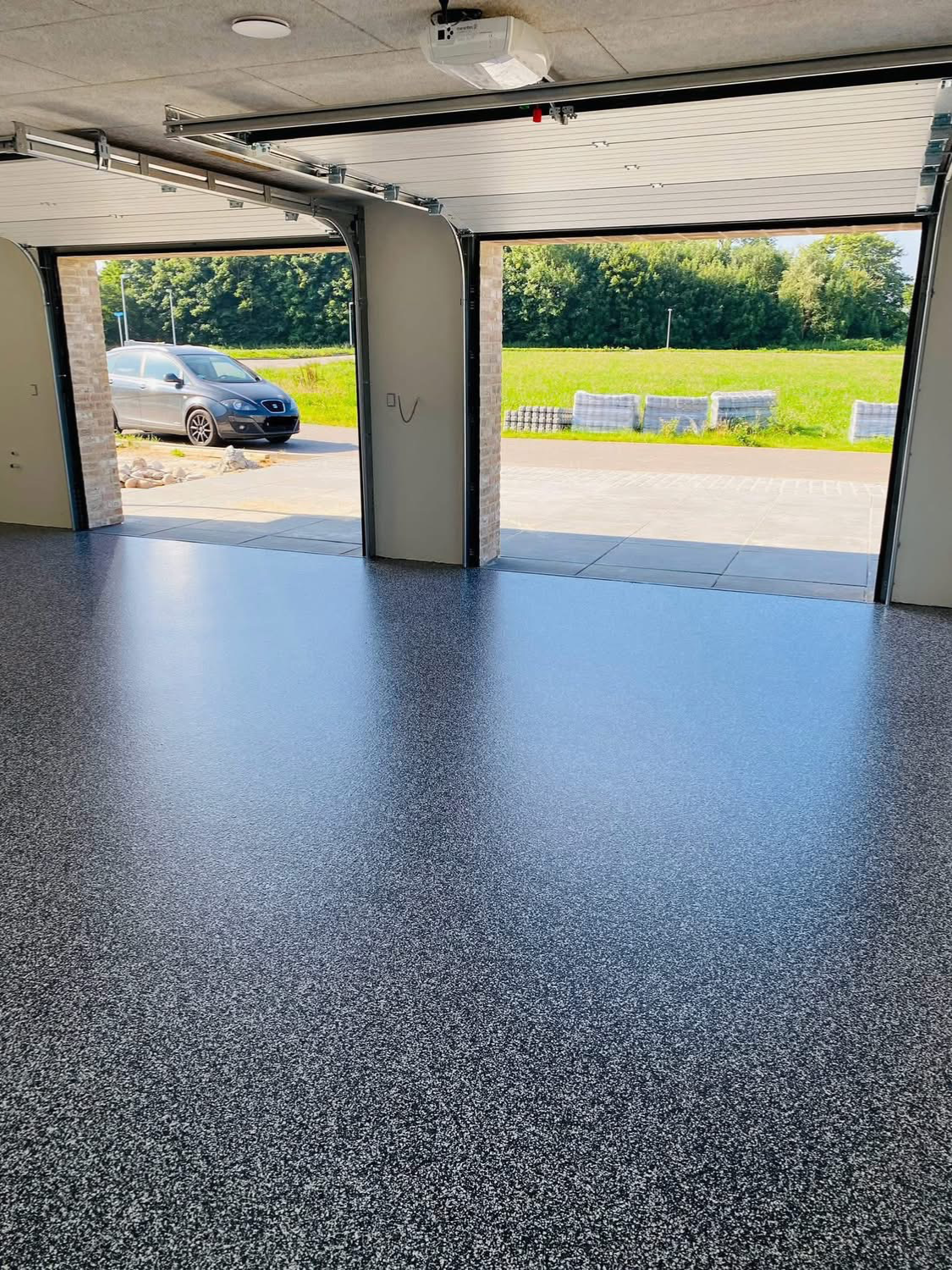 Gulv i en garage med åbne porte, der viser en bil og en grøn græsmark i baggrunden.