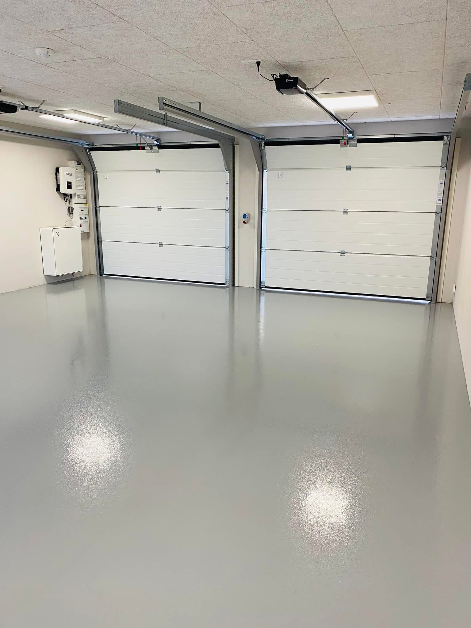 Lyst garageinteriør med to hvide garageporte og glat, grå gulv.