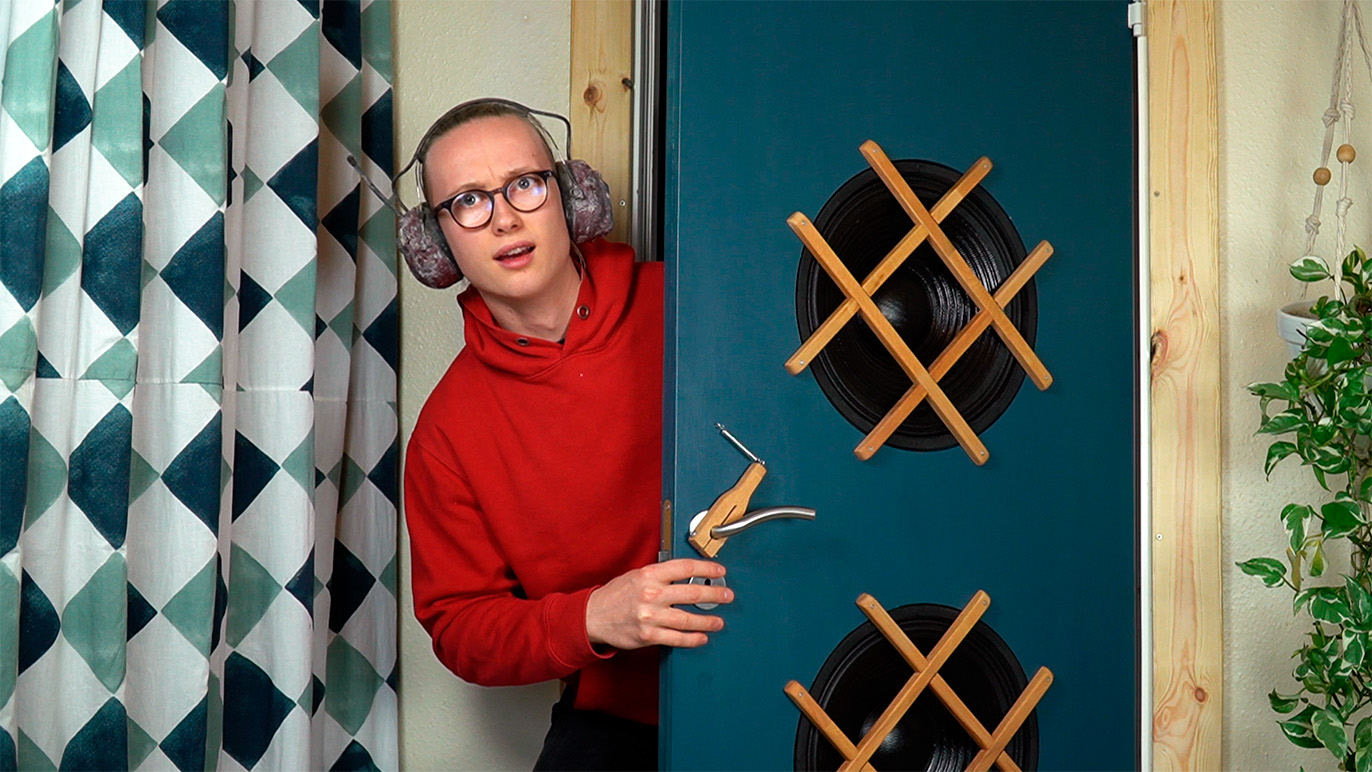 En ung mand i rød hættetrøje kigger nysgerrigt ud fra en dør med plader og træstænger.