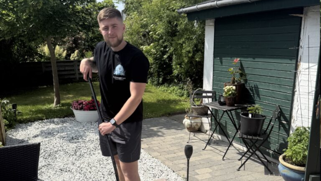 En ung mand står med en spade i haven, omgivet af planter og grus, iført sort t-shirt og shorts.