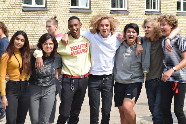 Gruppe af unge mennesker smilende og poserende sammen i en skolegård på en solrig dag.